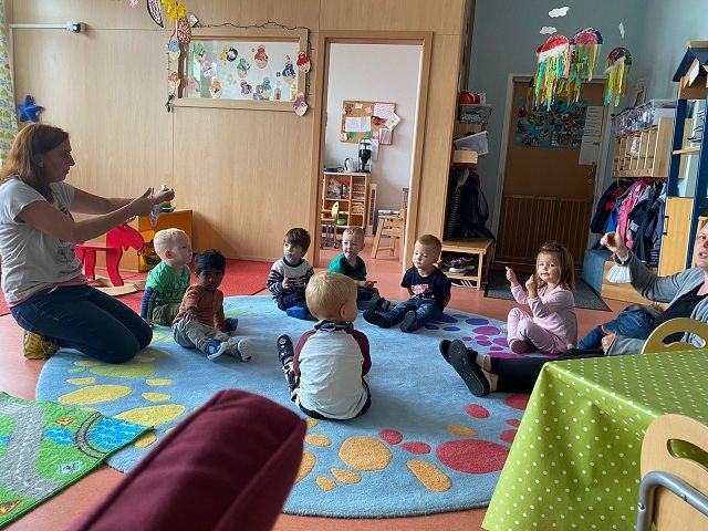 Kinder und Erzieherin sitzen gemeinsam im Sitzkreis auf dem Boden